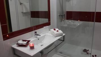 A modern bathroom with a glass shower enclosure, a white sink with a mirror above it, and a small table with a candle and a towel.