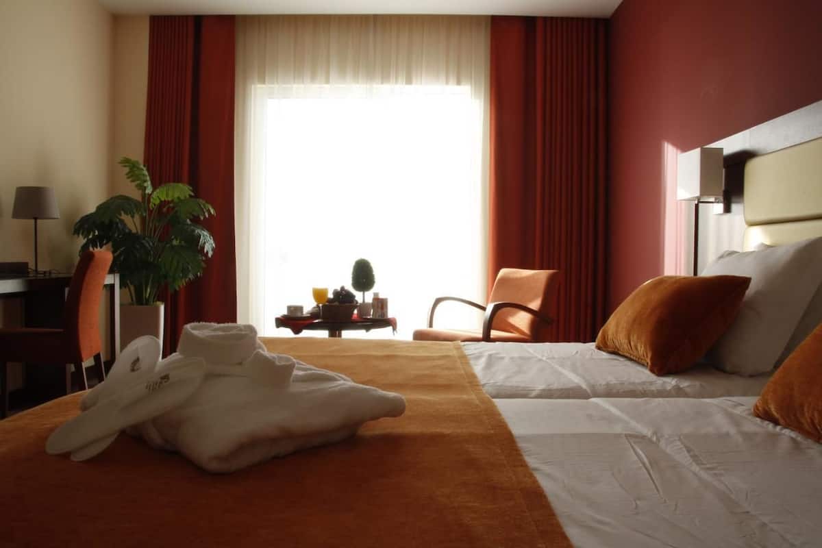 Ein Hotelzimmer mit zwei Betten, einem großen Fenster mit Vorhängen, einer Pflanze, einem Tisch mit Lampe und einem Sessel.