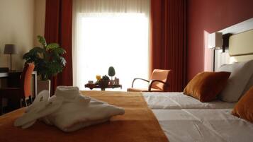 Quarto de hotel com duas camas, uma janela grande com cortinas, uma planta, uma mesa com abajur e uma cadeira.