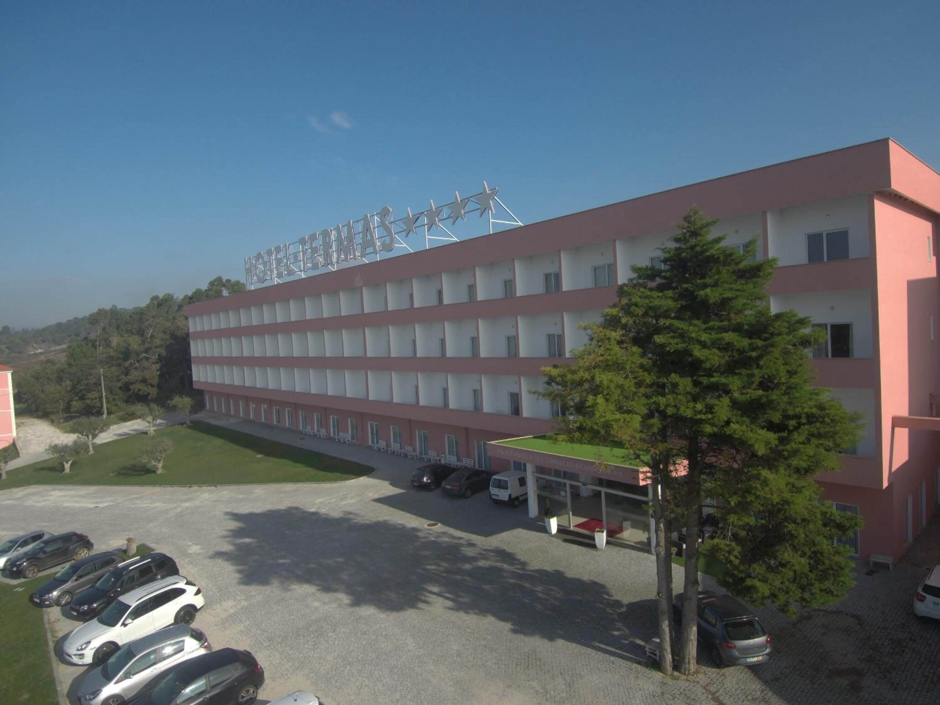 Un hotel de varios pisos con una fila de habitaciones en cada planta, un estacionamiento con varios autos y una pequeña área verde con árboles.