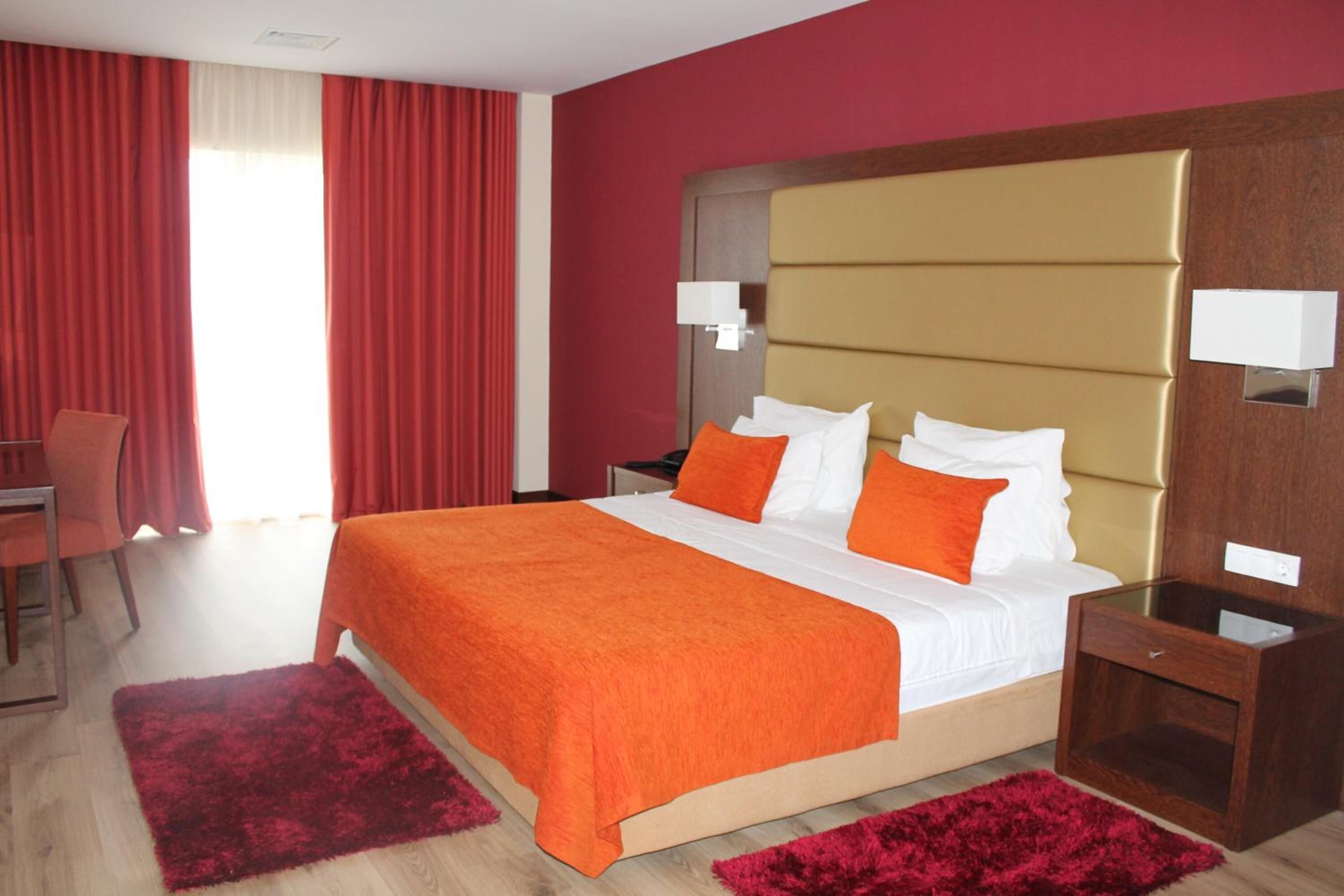 A hotel room with a large bed, red curtains, a wooden headboard, and a small bedside table.