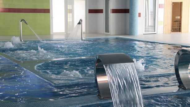 Une piscine intérieure dotée de jets d’eau, entourée de murs ornés de motifs à rayures rouges, et de portes donnant accès à d’autres espaces.