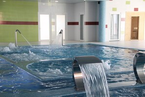 Une piscine intérieure dotée de jets d’eau, entourée de murs ornés de motifs à rayures rouges, et de portes donnant accès à d’autres espaces.