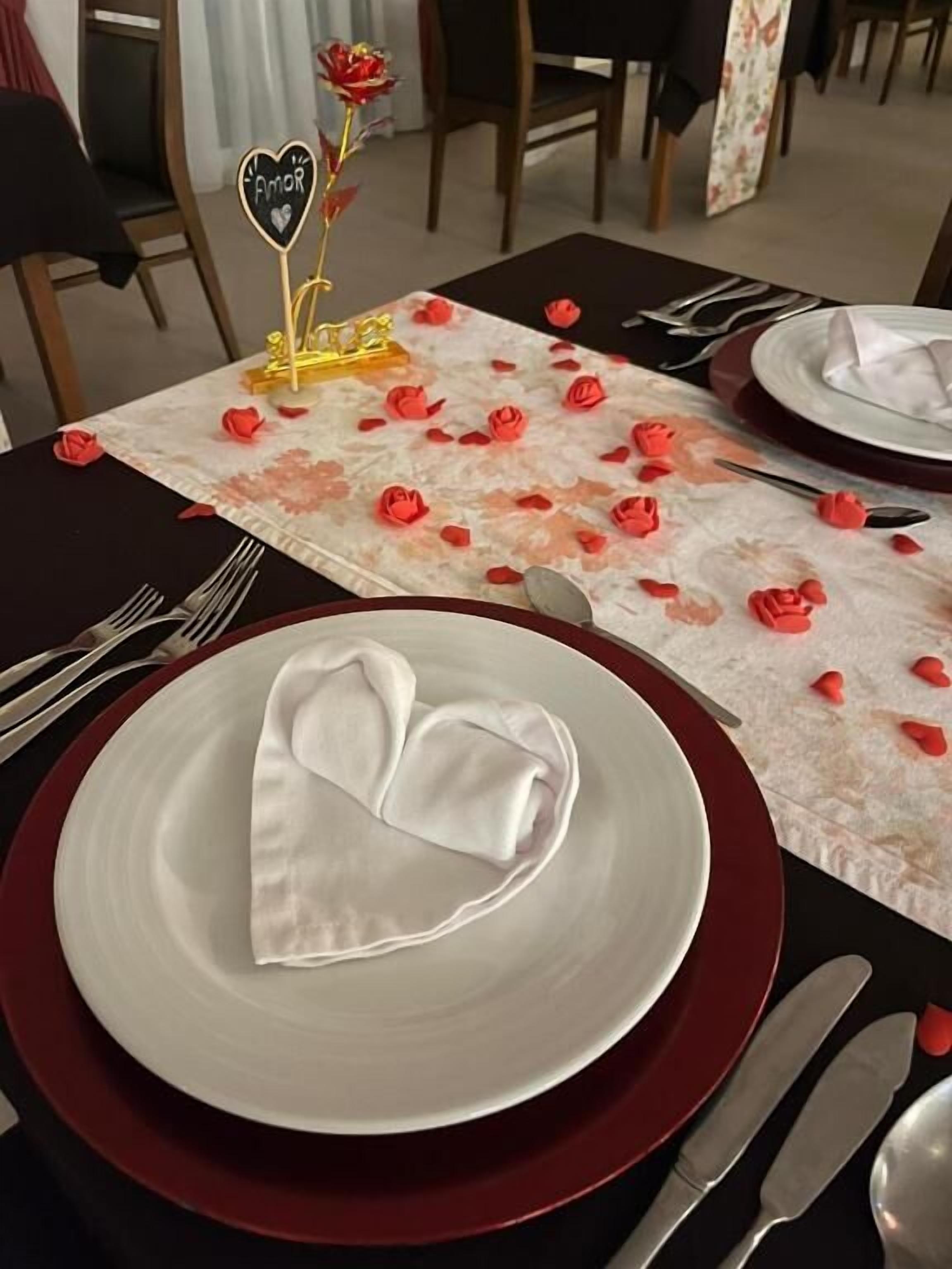 A dining table set with a heart-shaped napkin, red rose petals, and a heart-shaped napkin holder with the word 'Amor'.