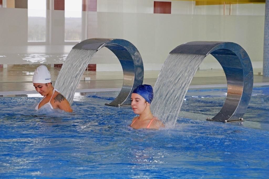 Two people in a swimming pool with water flowing from above.