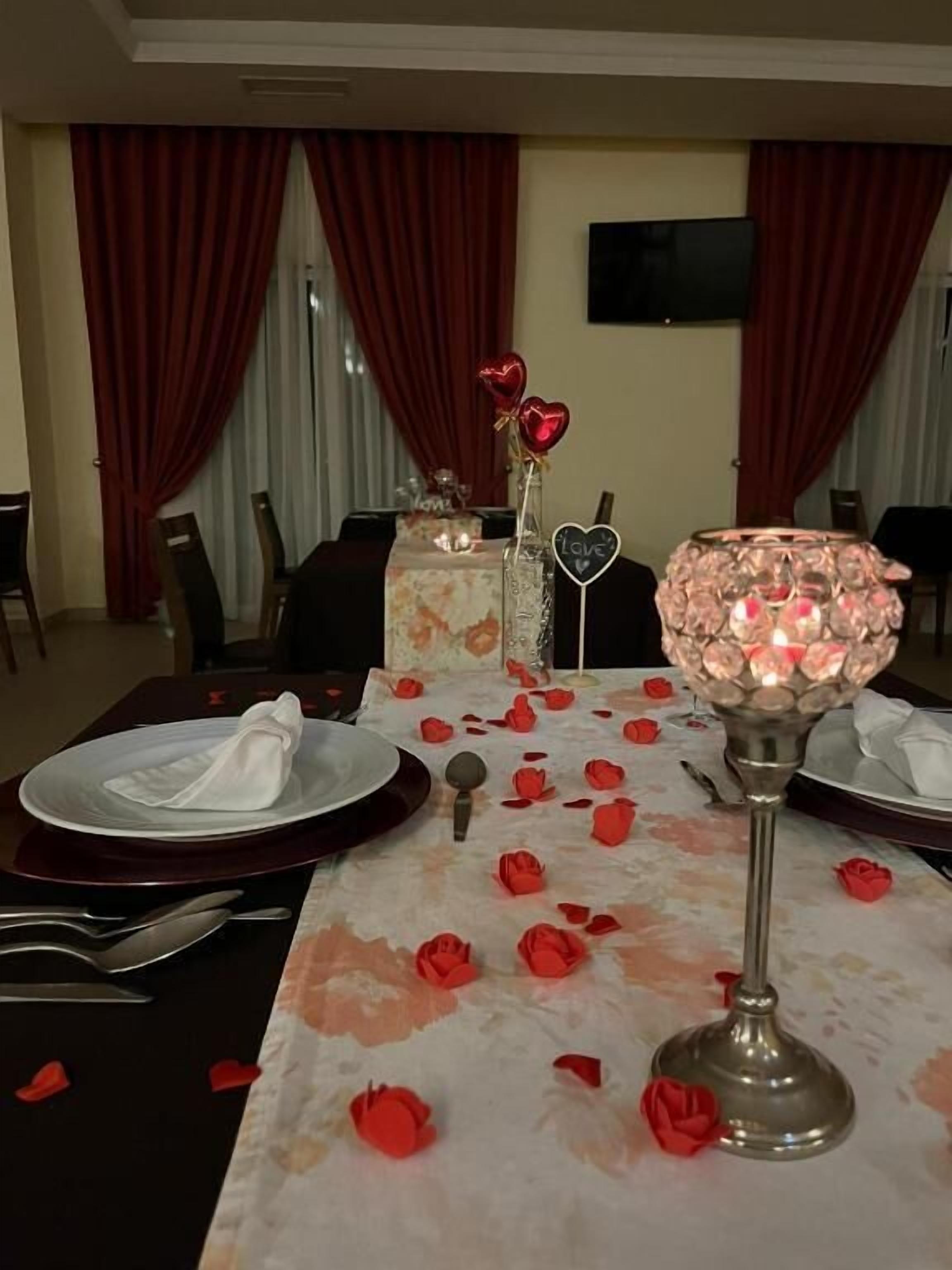 Una mesa de comedor adornada con un mantel floral, pétalos de rosa roja esparcidos sobre la superficie y un centro de mesa con velas.