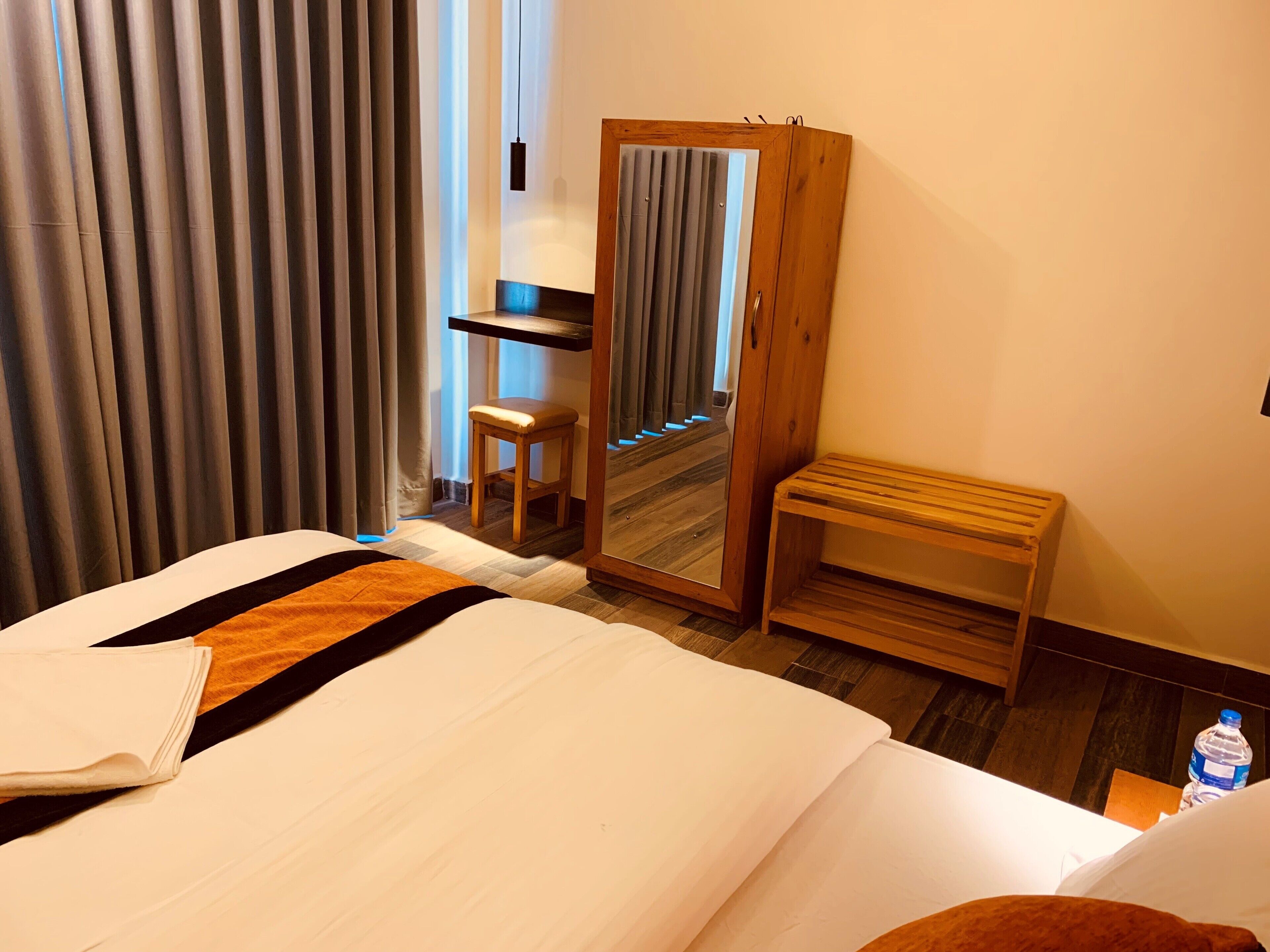 A hotel room with a bed, a wooden bench, a small stool, a mirror, and a water bottle on the floor.