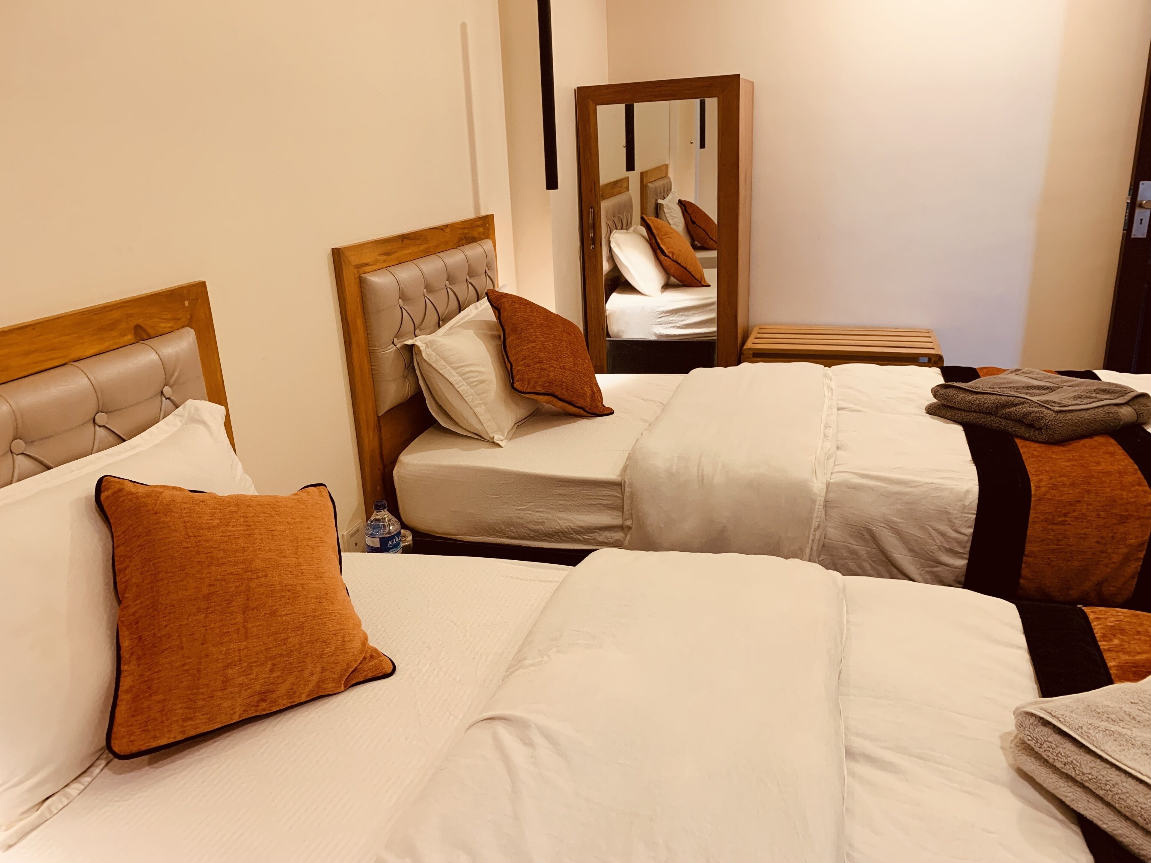 A hotel room with two beds, each with a headboard, white bedding, and brown pillows. A wooden nightstand with a lamp is between the beds.