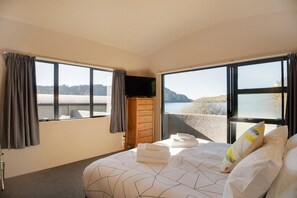 A hotel room with a bed, a flat-screen TV, a wooden dresser, and large windows offering a view of the sea.