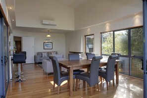 A spacious living area with a wooden dining table, grey upholstered chairs, a grey sofa, and a large window providing a view of the outdoors.