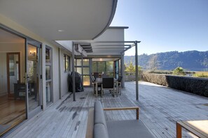 Un espacio al aire libre techado con mesa y sillas, un sofá, y vistas a un lago y colinas.