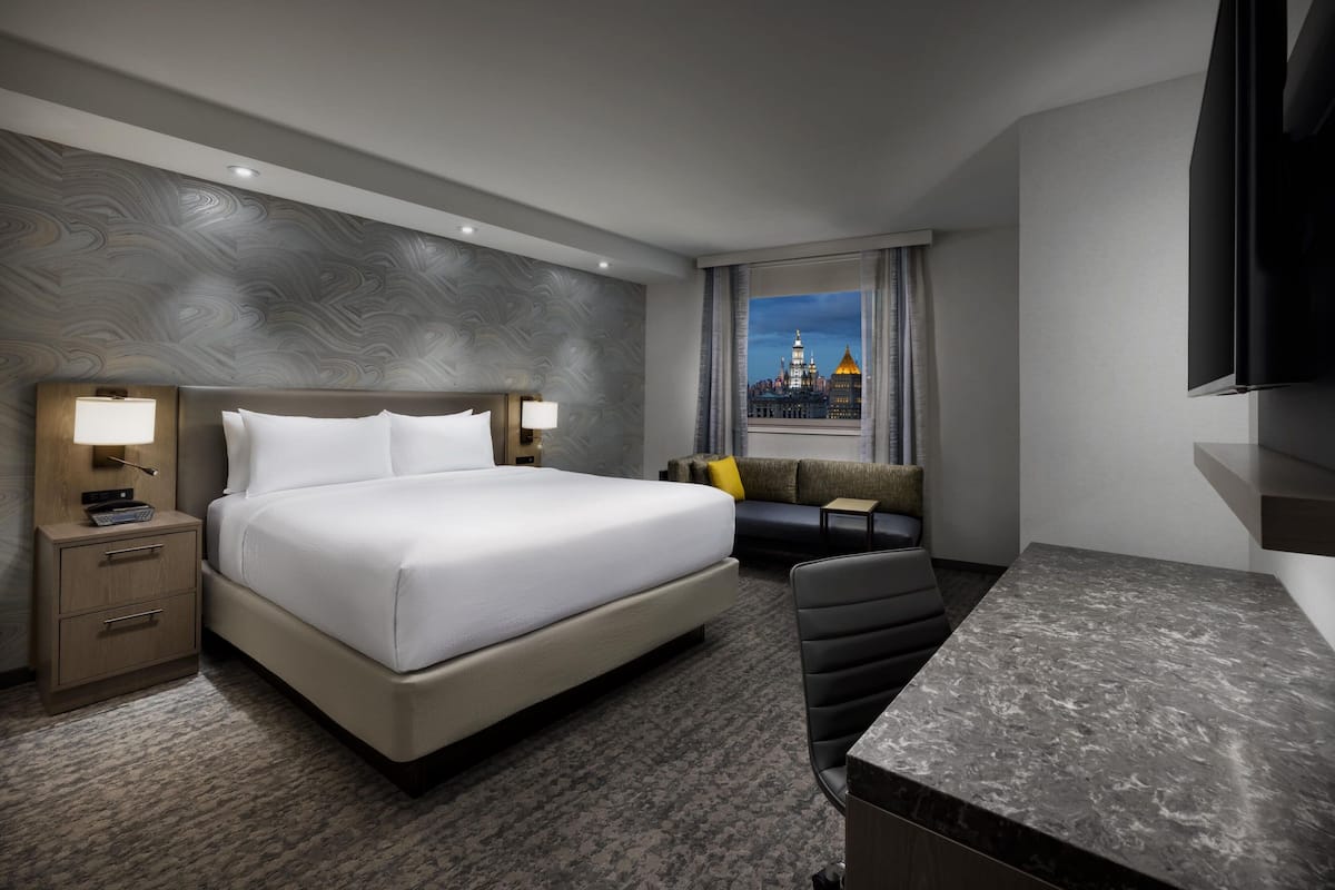A hotel room with a large bed, a desk, a chair, a nightstand, and a view of a cityscape through the window.