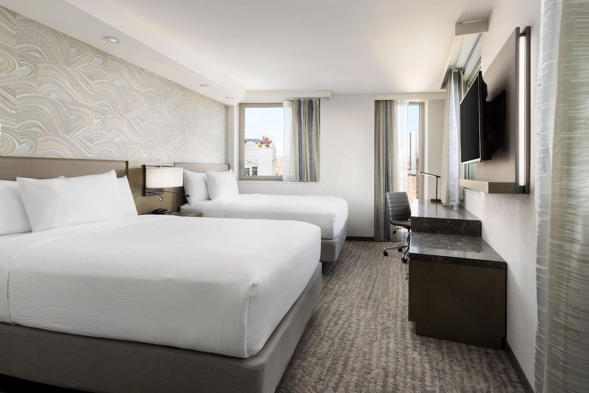 A hotel room with two beds, a desk, a flat-screen TV, and a window with curtains.