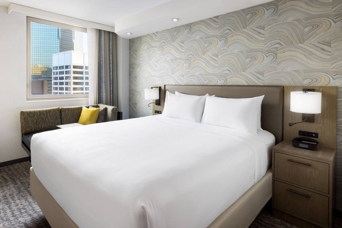 A hotel room with a large bed, a bedside table with a lamp, a window with a view of a building, and a patterned wall.