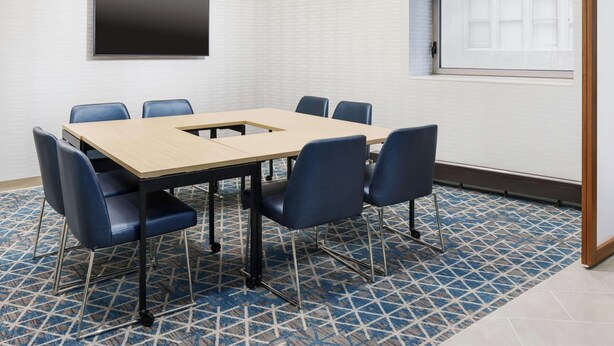 Un salón de conferencias con una mesa rectangular, seis sillas azules, un televisor de pantalla plana montado en la pared y una alfombra estampada.