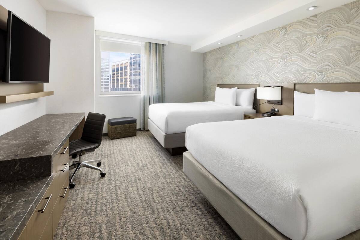 A hotel room with a large bed, a desk with a chair, a TV mounted on the wall, and a window with a view of buildings.