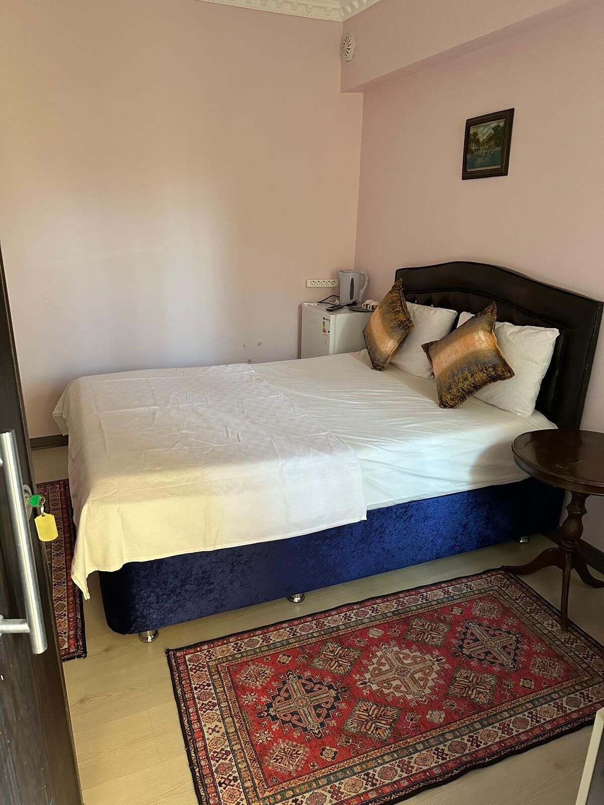 Un dormitorio con una cama, una mesita de noche pequeña, una lámpara de noche, un tapete rojo estampado y un cuadro colgado en la pared.