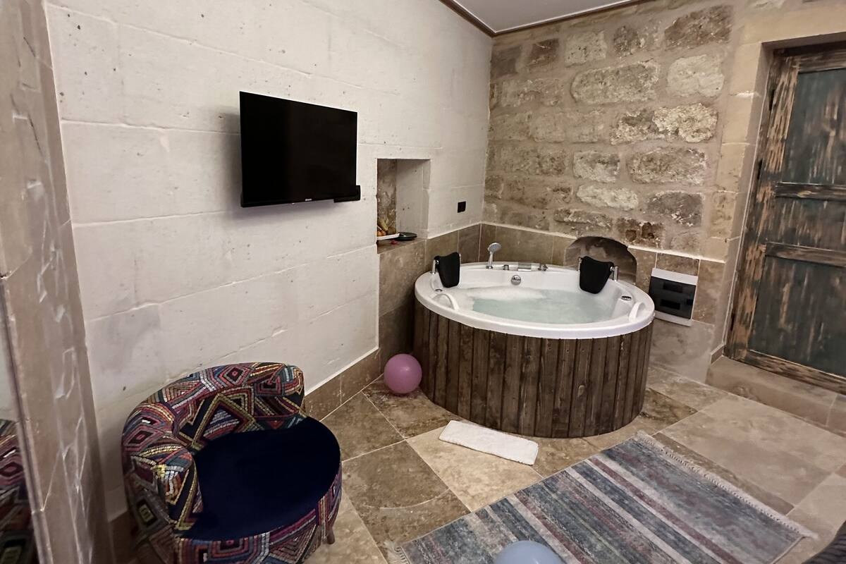 Un baño con pared de piedra, una bañera de madera, un televisor montado en la pared y una silla estampada.