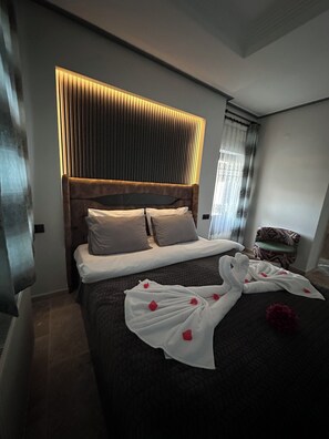 A modern hotel room with a large bed, a headboard with vertical slats, and a bedside table with a patterned cushion. - DİRİLİŞ BANARYA PANSİYON (Ürgüp)