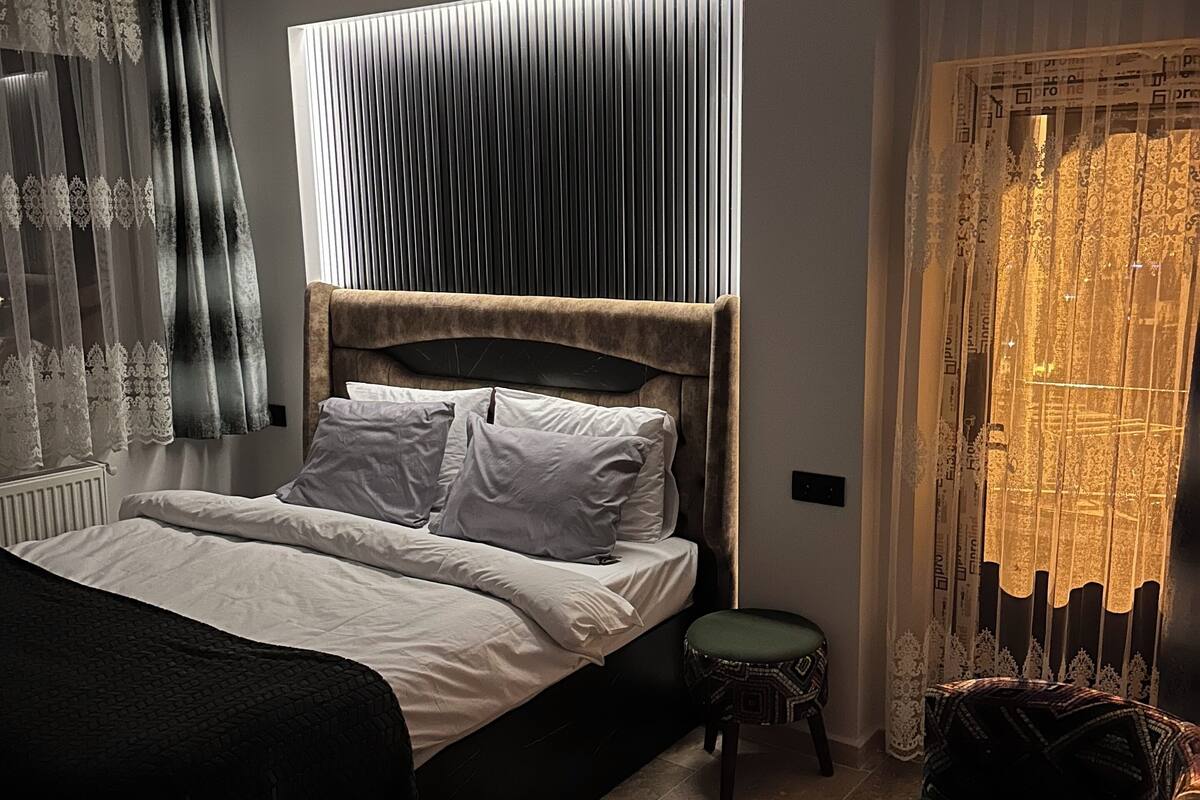 Un dormitorio moderno con una cama grande, un cabecero con líneas verticales, un taburete pequeño y una silla.