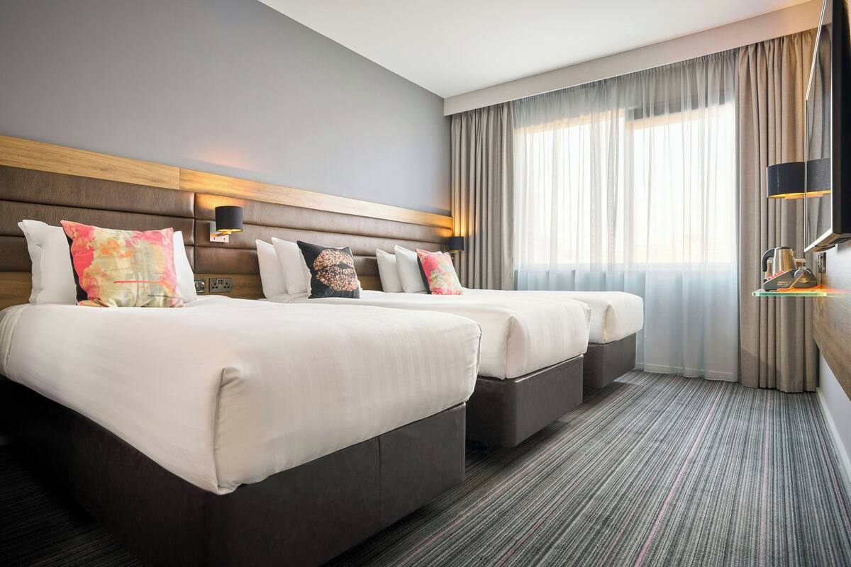 A hotel room with two beds, a headboard, a bedside table, a lamp, and a view of the outdoors through the window.