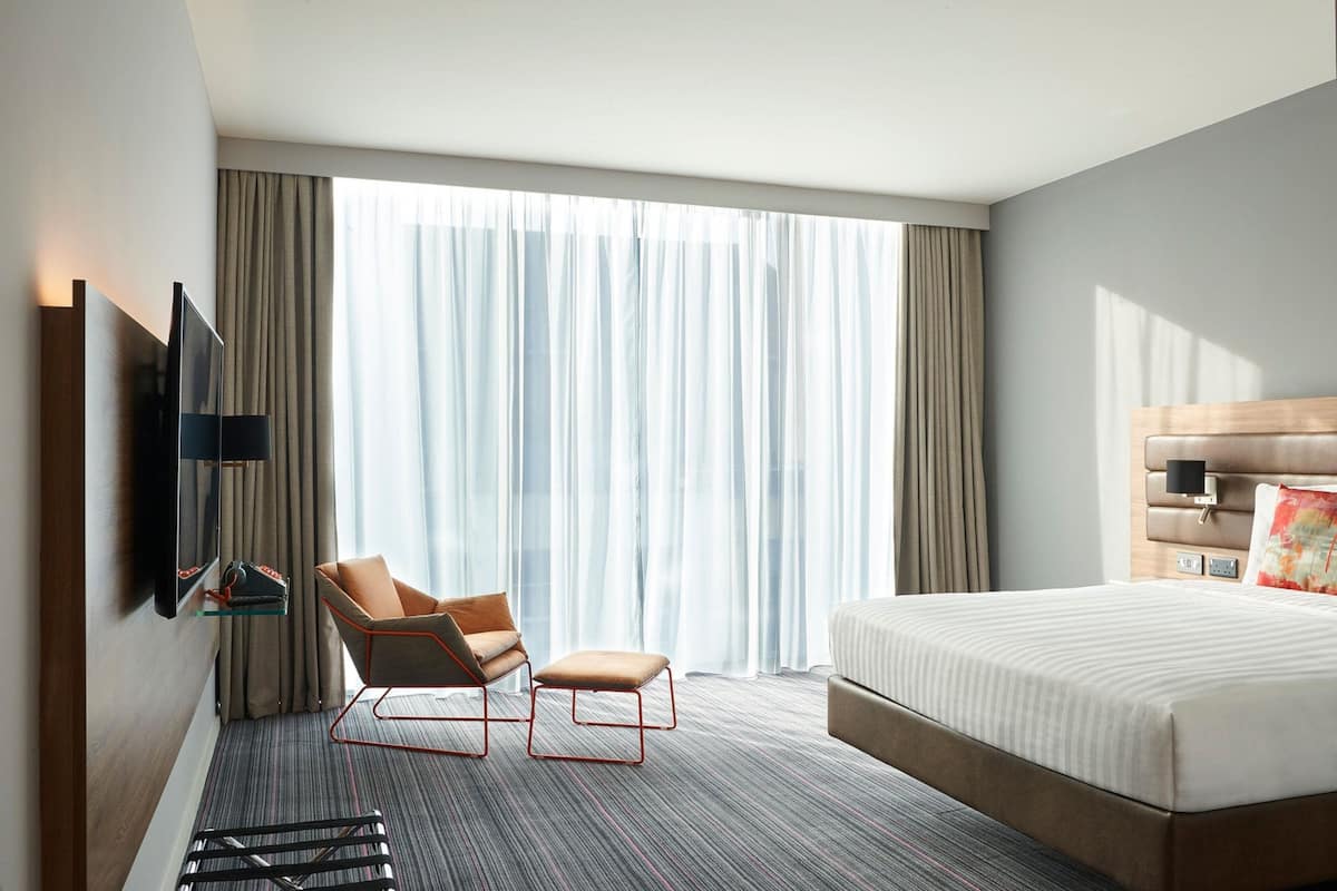 A hotel room with a large bed, a chair, a TV mounted on the wall, and a window with curtains.