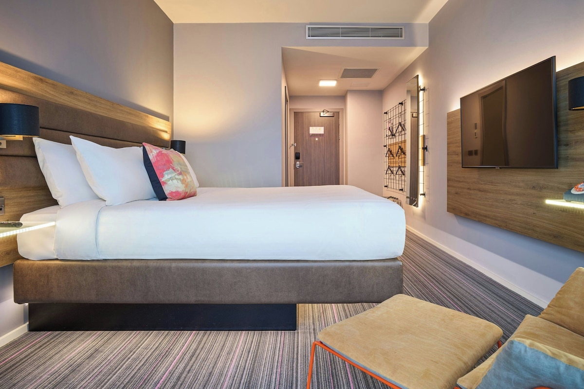 A modern hotel room with a large bed, a flat-screen TV mounted on the wall, and a small seating area with a cushioned bench.