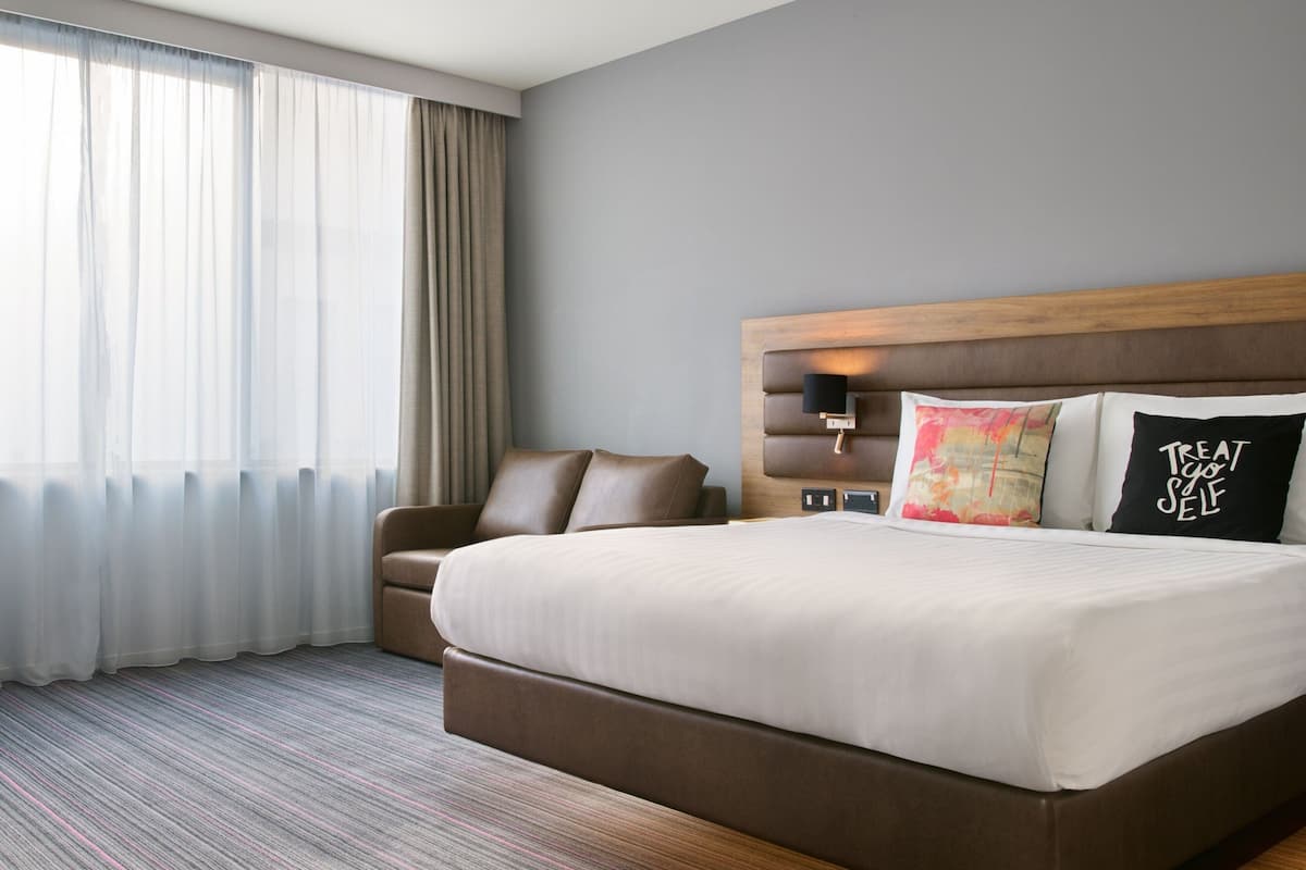 A hotel room with a large bed, a chair, a nightstand, and a window with curtains.