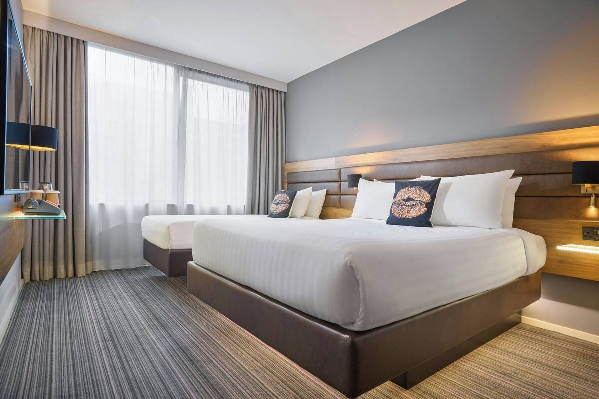 A hotel room with a large bed, a smaller bed, a desk with a lamp, and a view of the outdoors through large windows.
