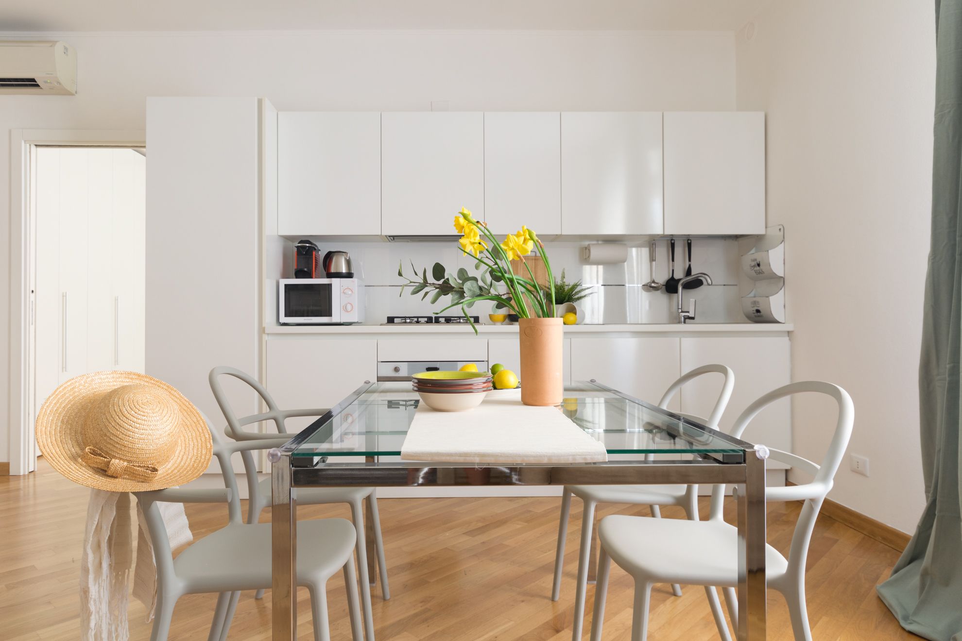 A modern kitchen with white cabinets, a glass dining table, and a vase with yellow flowers.