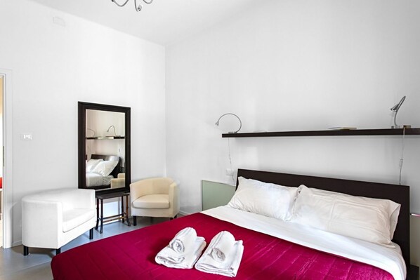 A hotel room with a large bed, a bedside table, a chair, a mirror, and a shelf.