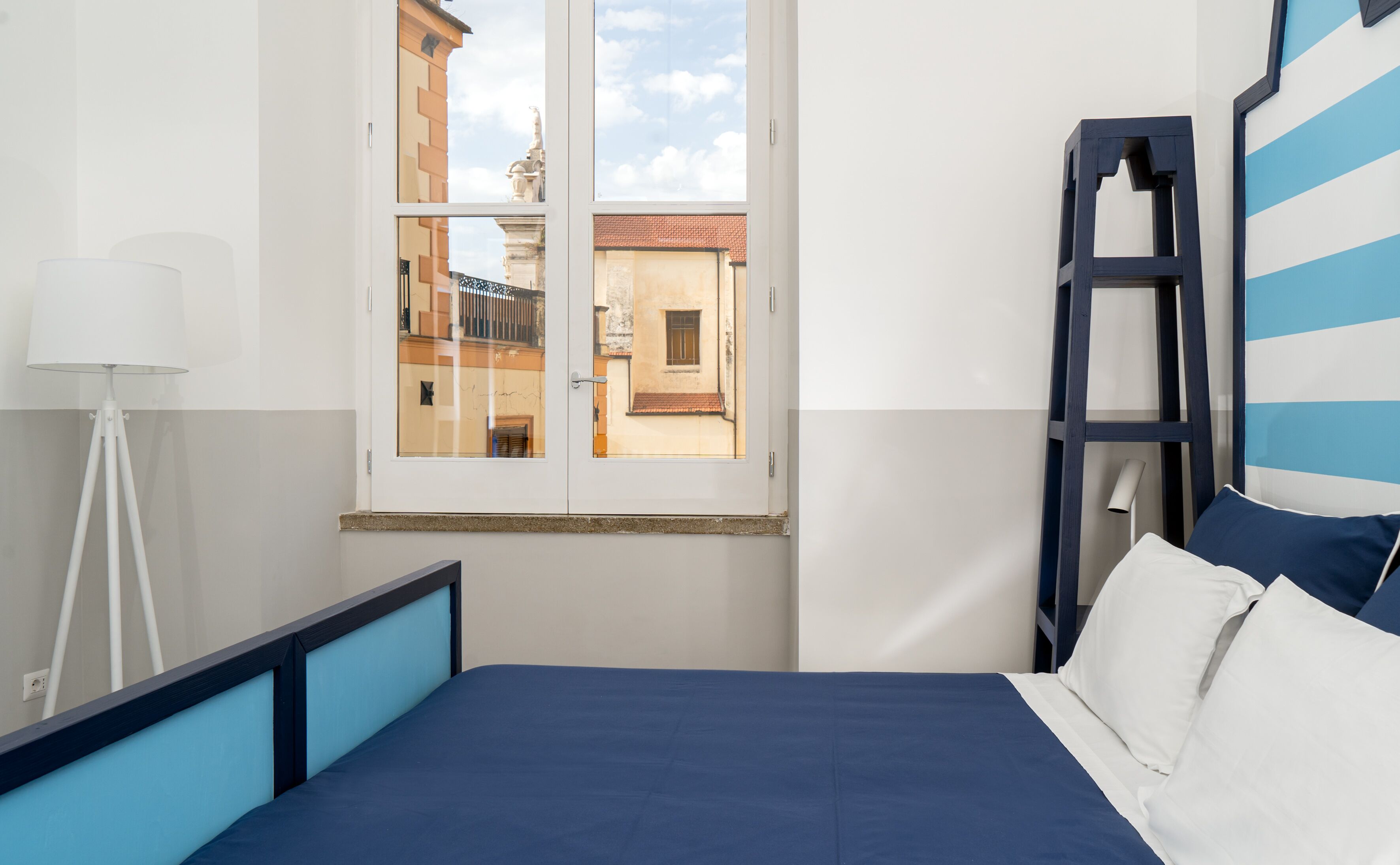 A bedroom with a bed, a window with a view of buildings, a floor lamp, and a ladder against the wall.