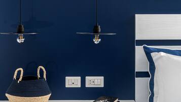 Un dormitorio con una pared azul oscuro, una mesita de noche blanca, una cama azul con almohadas blancas y dos lámparas colgantes.