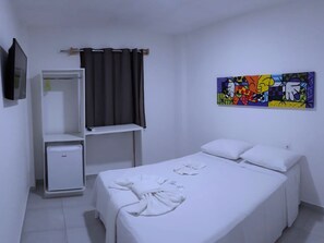 A hotel room with a bed, a wardrobe, a small fridge, a TV, and a colorful painting on the wall. - Jardins Premium (Ipojuca)