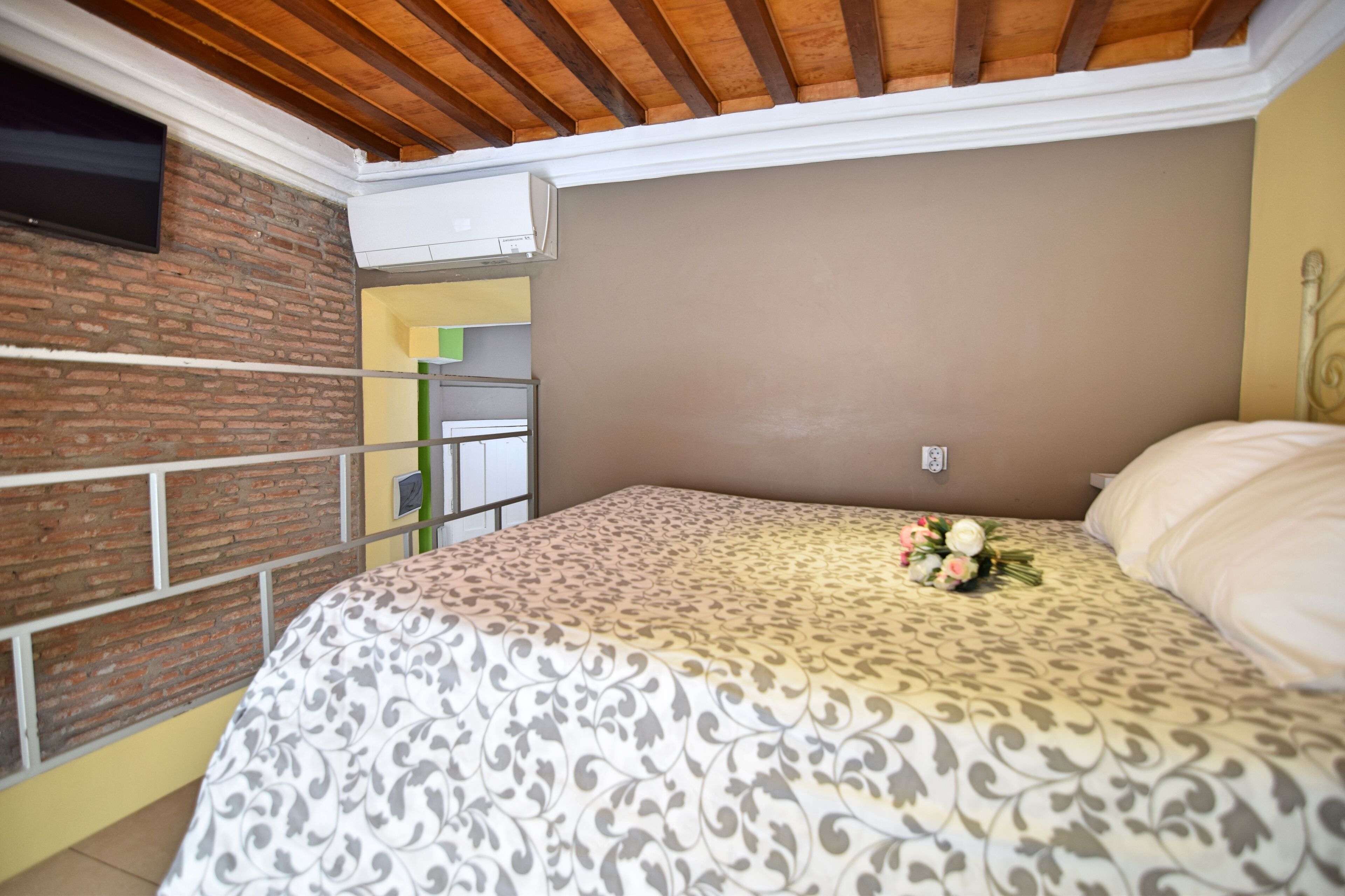Une chambre à coucher avec une couvre-lit à motifs, un bouquet de fleurs, un téléviseur fixé à un mur en briques et un appareil de climatisation.
