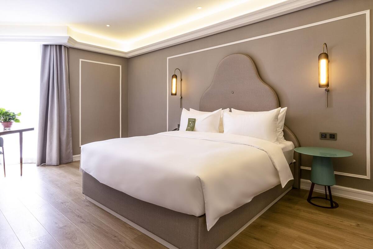 A hotel room with a large bed, bedside tables, a small round table, and a potted plant.