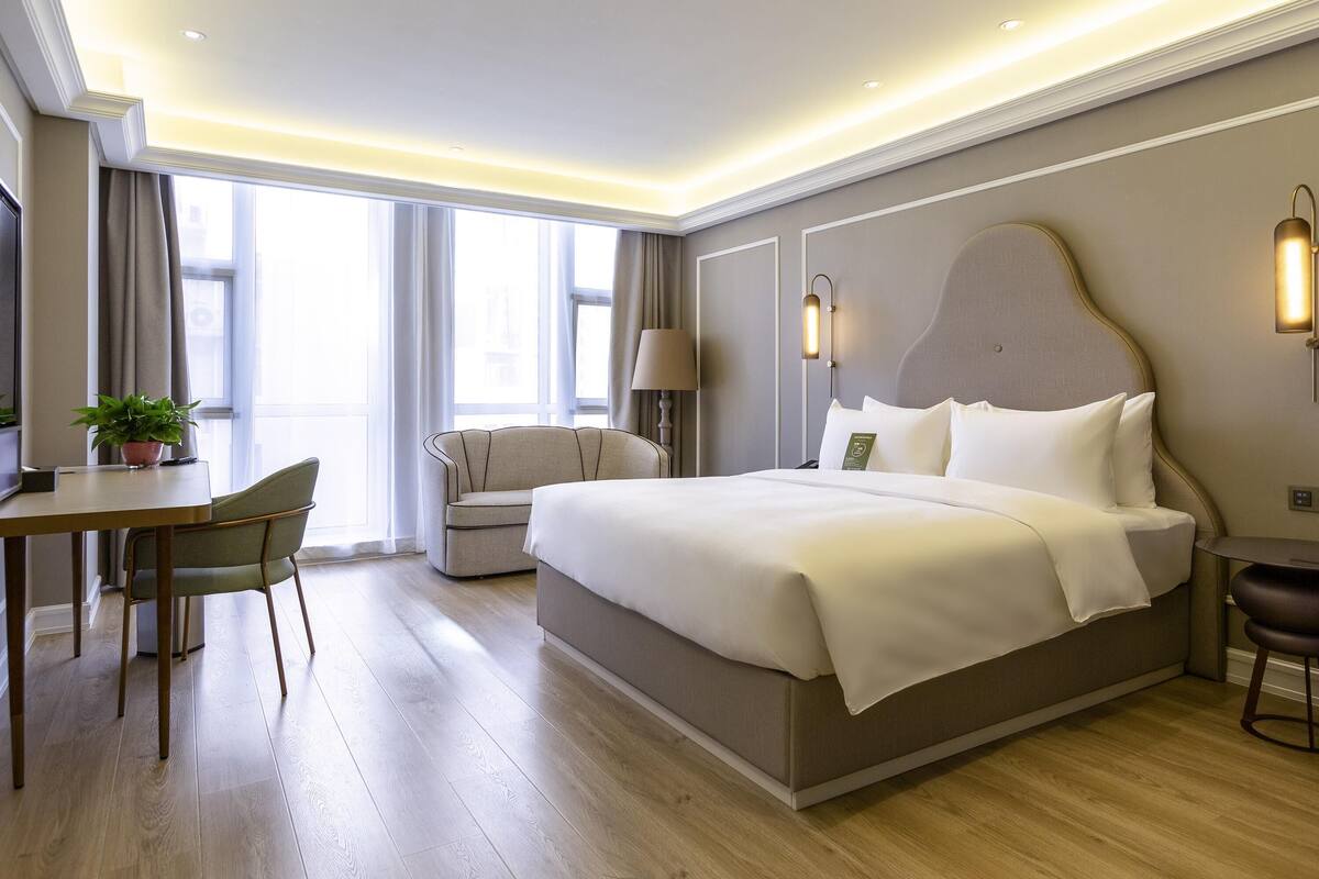 A modern hotel room with a large bed, a desk, a chair, and a potted plant.