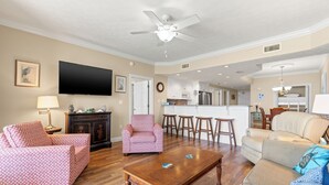 A living room with a ceiling fan, a TV, a sofa, a coffee table, and a kitchen area in the background. - Treasure Island 1007 (Panama City Beach)