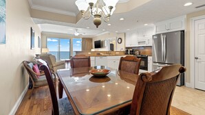 A spacious dining area with a glass-top table, wooden chairs, a chandelier, and a kitchen in the background. - Treasure Island 1007 (Panama City Beach)
