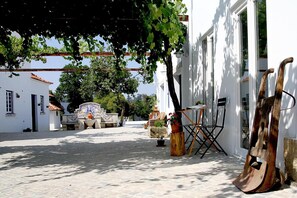 Una calle adoquinada con edificios blancos, un árbol que da sombra y una escultura de madera rústica.