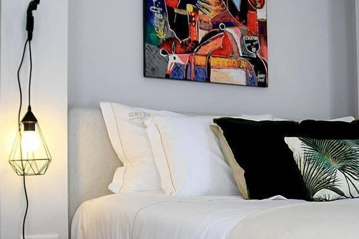 Un dormitorio moderno con cama, mesita de noche de madera, lámpara de pie y un cuadro abstracto de colores en la pared.