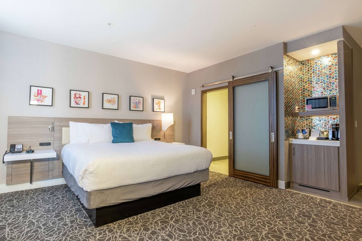 A hotel room with a bed, bedside table, wall art, a wardrobe, and a kitchenette area.