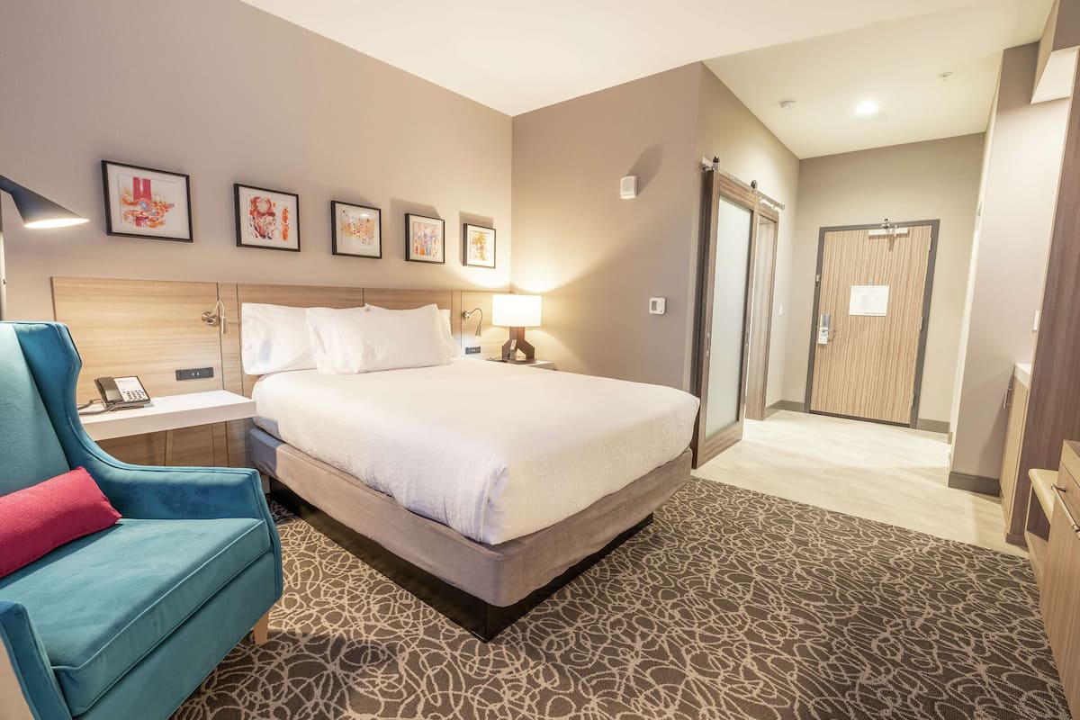 A hotel room with a bed, a desk with a phone, a chair with a red pillow, and a shower area.