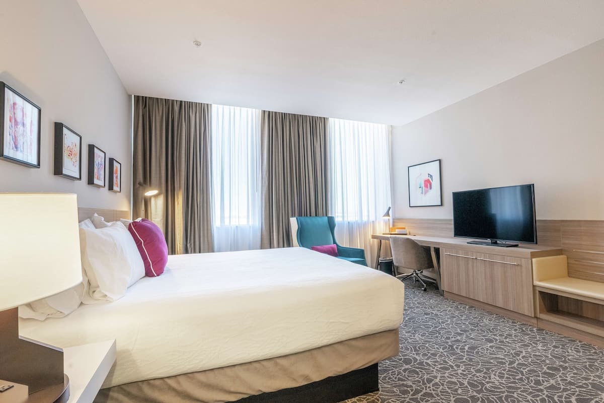 A hotel room with a large bed, a desk with a TV, a chair, and a desk lamp.