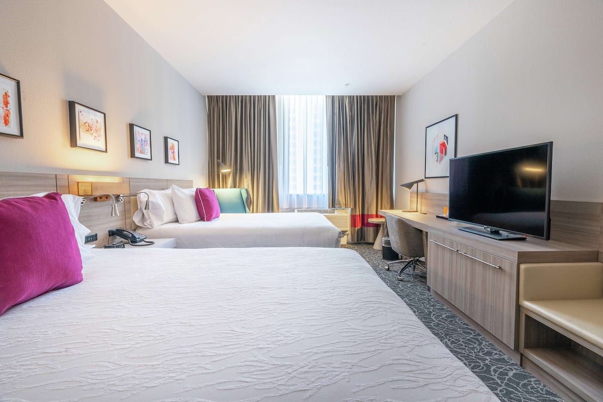 A hotel room with two beds, a TV, a desk, and a window with curtains.