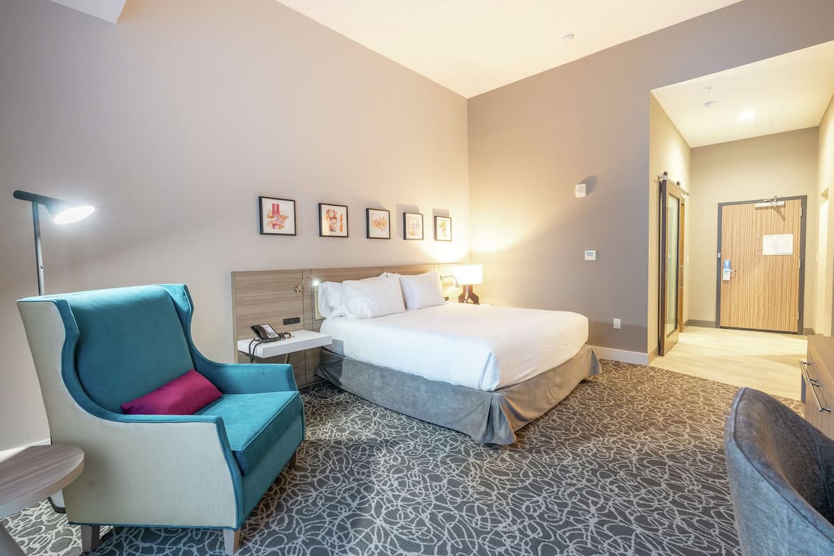 A hotel room with a bed, a desk, a chair, and a lamp. There are framed pictures on the wall and a door leading to another room.