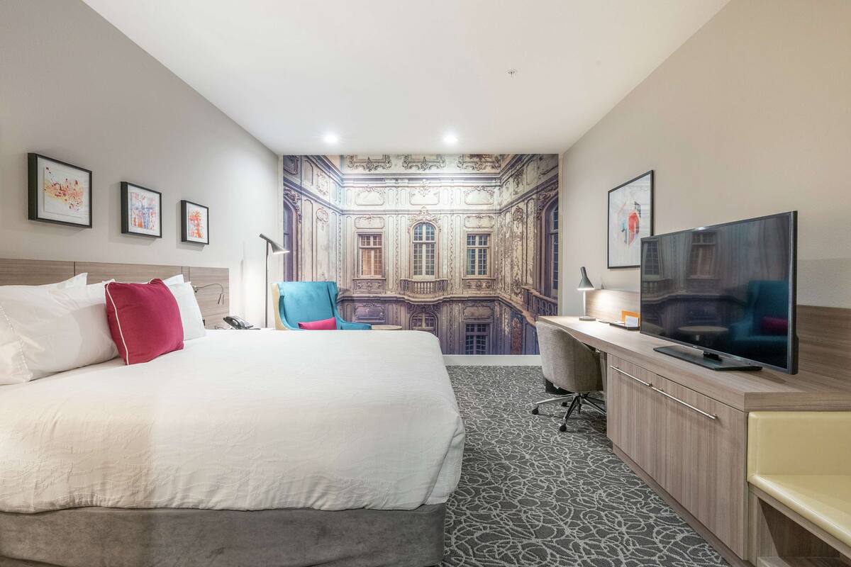 A hotel room with a large bed, a desk with a TV, and a chair. There are framed pictures on the wall and a mural of a building.
