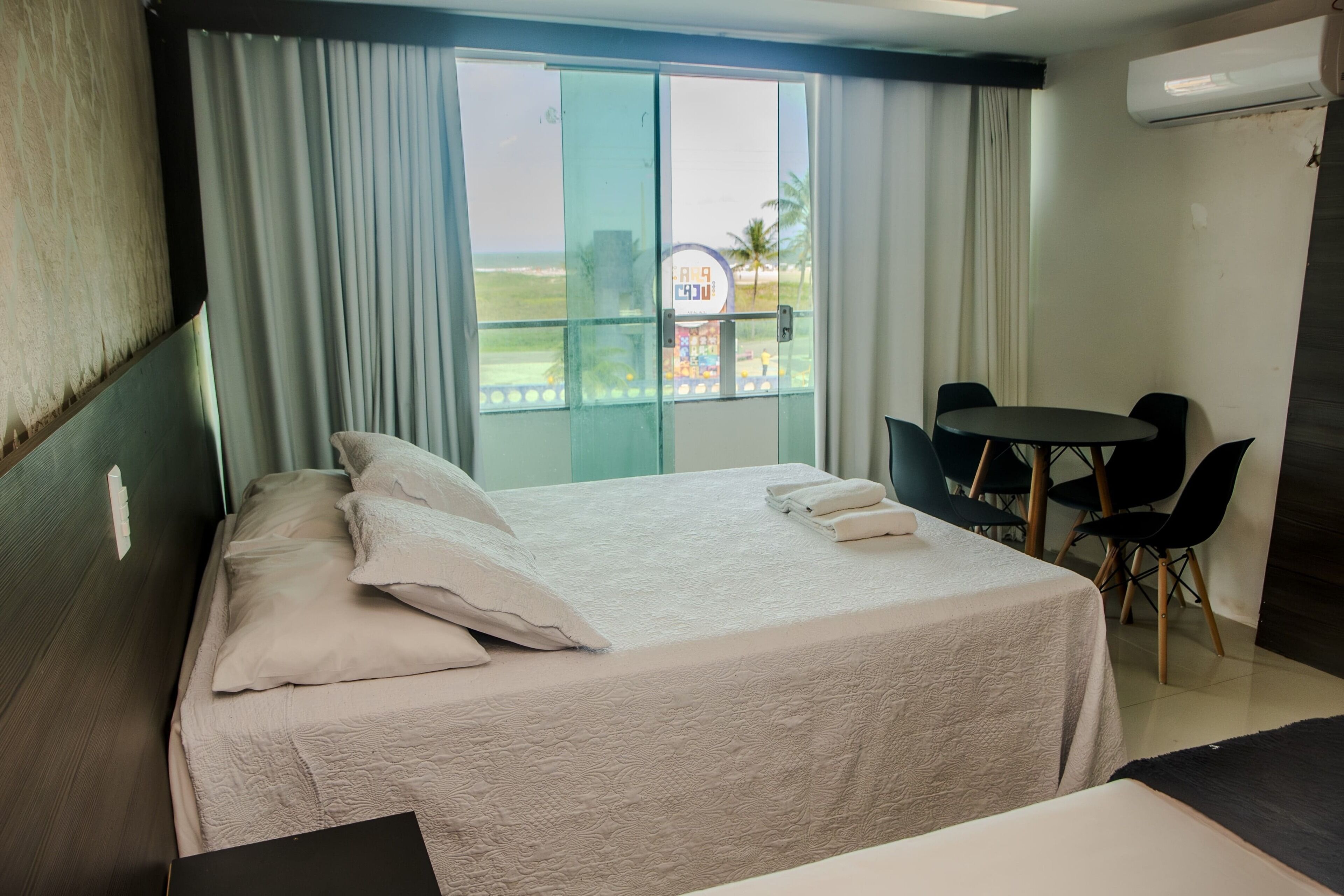 Habitación de hotel con cama, una mesita, sillas, un balcón con vista a la playa y un equipo de aire acondicionado.