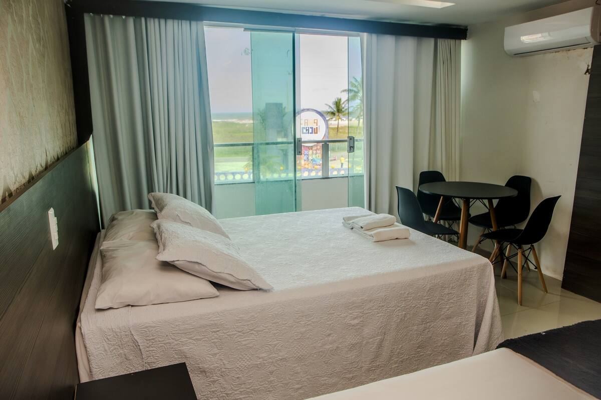 A hotel room with a bed, a small table, chairs, a balcony with a view of the beach, and an air conditioning unit.