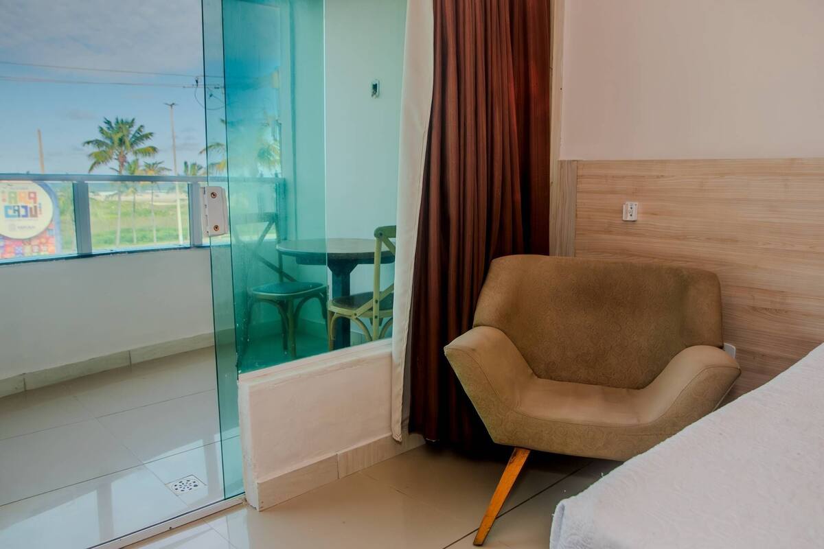 A hotel room with a bed, a brown armchair, a glass partition, and a view of palm trees and a signboard outside.